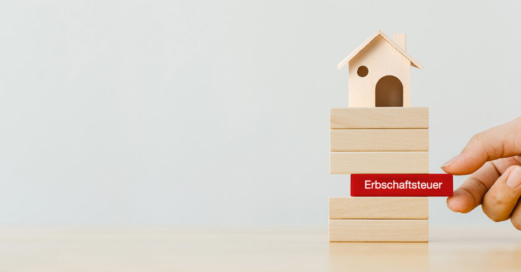 Holzhäuschen steht auf Holzklötzchen auf einem Tisch, ein rotes Holzklötzchen mit der Aufschrift Erbschaftsteuer wird von einer Hand herausgezogen | Erbschaftsteuer