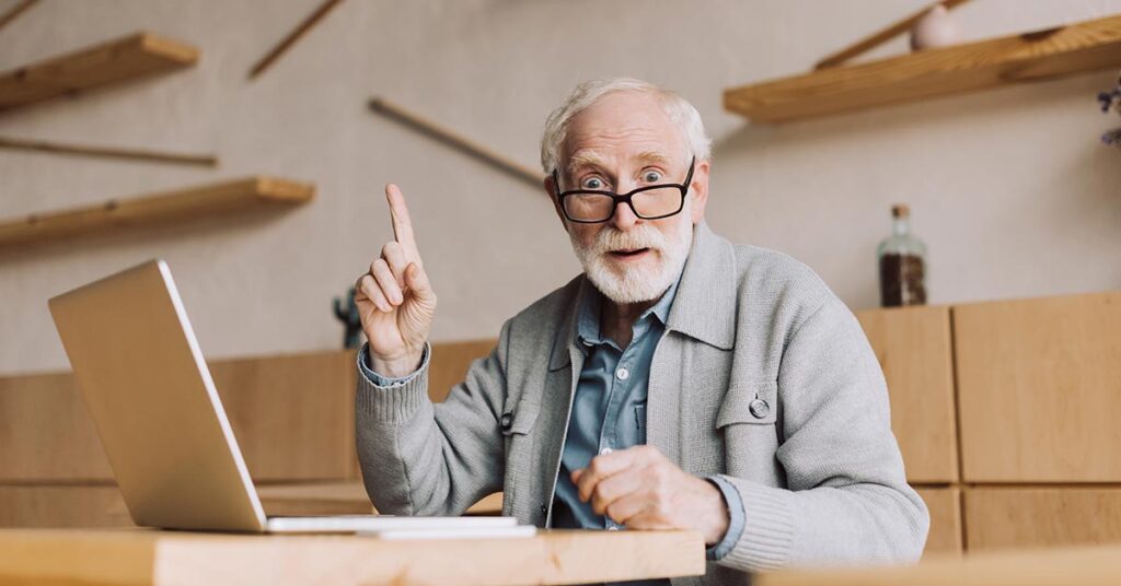Alter Mann sitzt im Café am Laptop, surft im Internet und hebt den Zeigefinger und macht große Augen, weil er eine Idee hat.