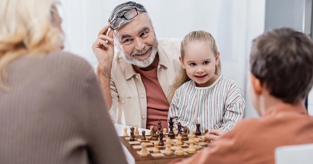 Ein Seniorenpärchen sitzt mit seinen Enkelkindern glücklich im Wohnzimmer und spielt Schach als Symbolbild für strategisch kluge Entscheidungen bei Investments für die Altersvorsorge | Wohnen im Alter